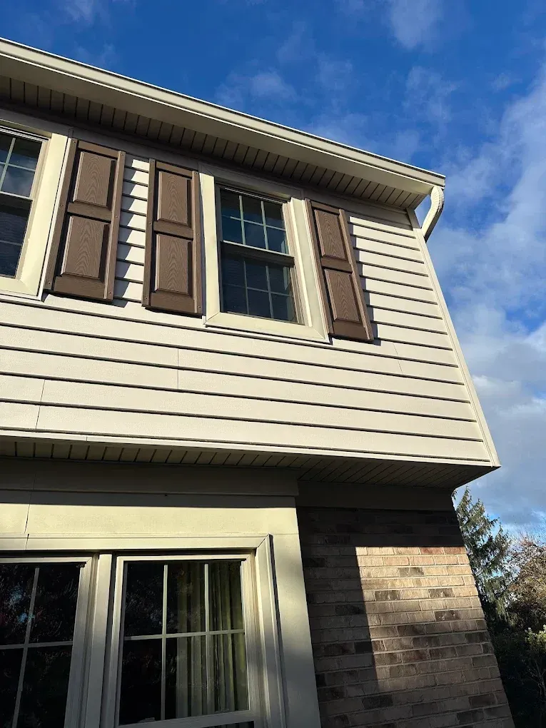 Exterior of a beige house with brown shutters, windows, and a brick corner under a blue sky