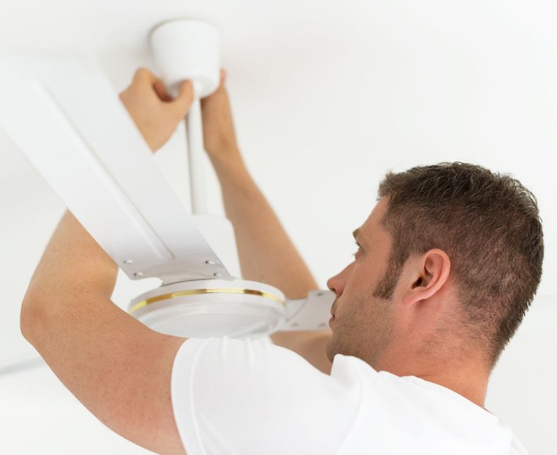 A Man In A White Shirt Is Installing A Ceiling Fan — Steve Mason Electrical In Port Macquarie, NSW