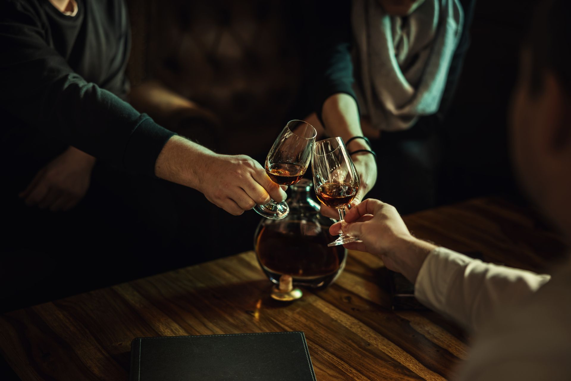 Persone che brindano con bicchieri di liquore color ambra versati in una caraffa su un tavolo di legno.