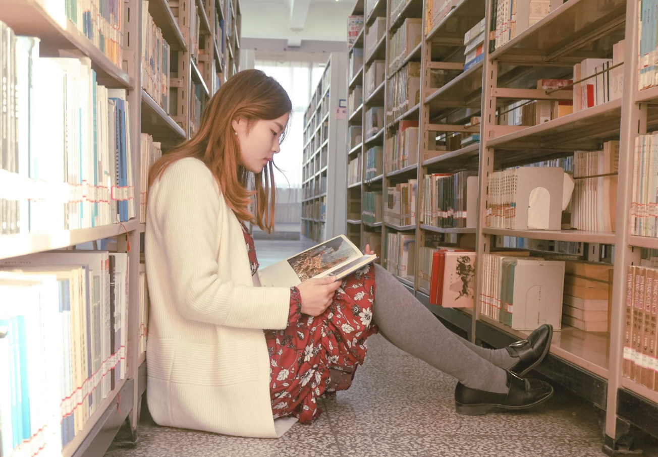 A woman is sitting on the floor in a library reading a book.