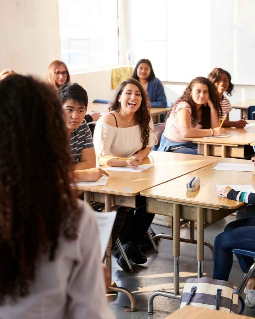 Students in a classroom. Some smiling and looking toward a person in front. Desks, window, bright lighting.