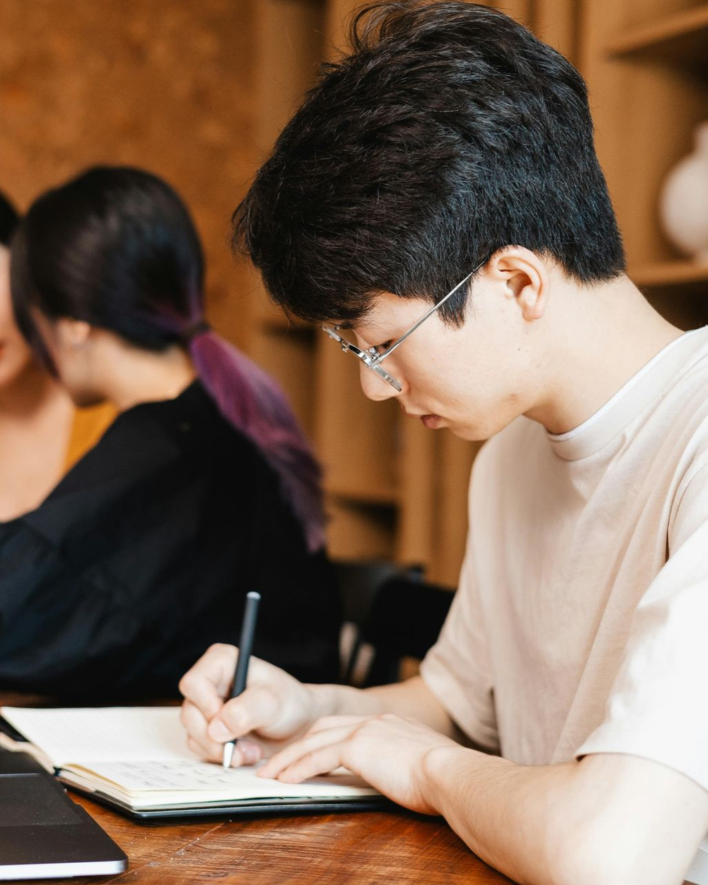 Students in a classroom. Some smiling and looking toward a person in front. Desks, window, bright lighting.