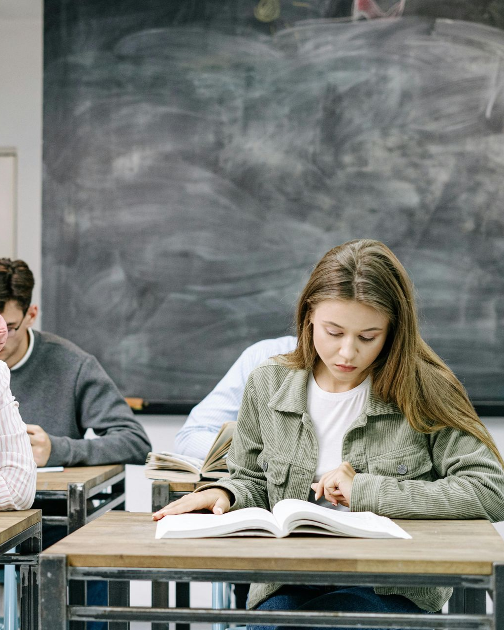Students in a classroom. Some smiling and looking toward a person in front. Desks, window, bright lighting.