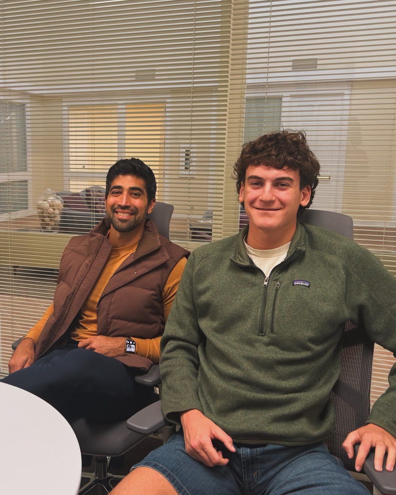 Two people seated in an office lounge, smiling beside a round table and venetian blinds.