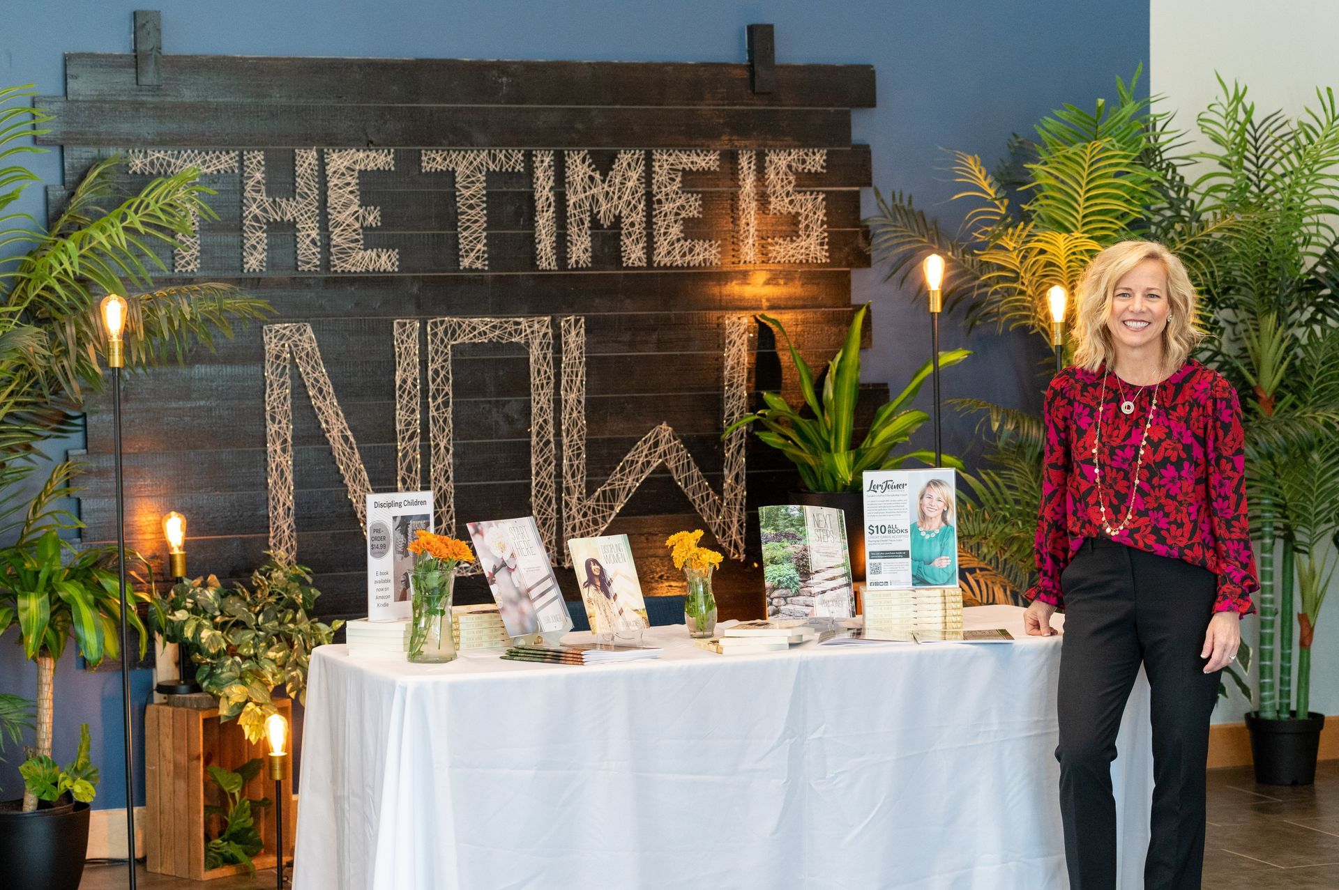 A woman is standing in front of a table with a sign that says `` the time is now ''.