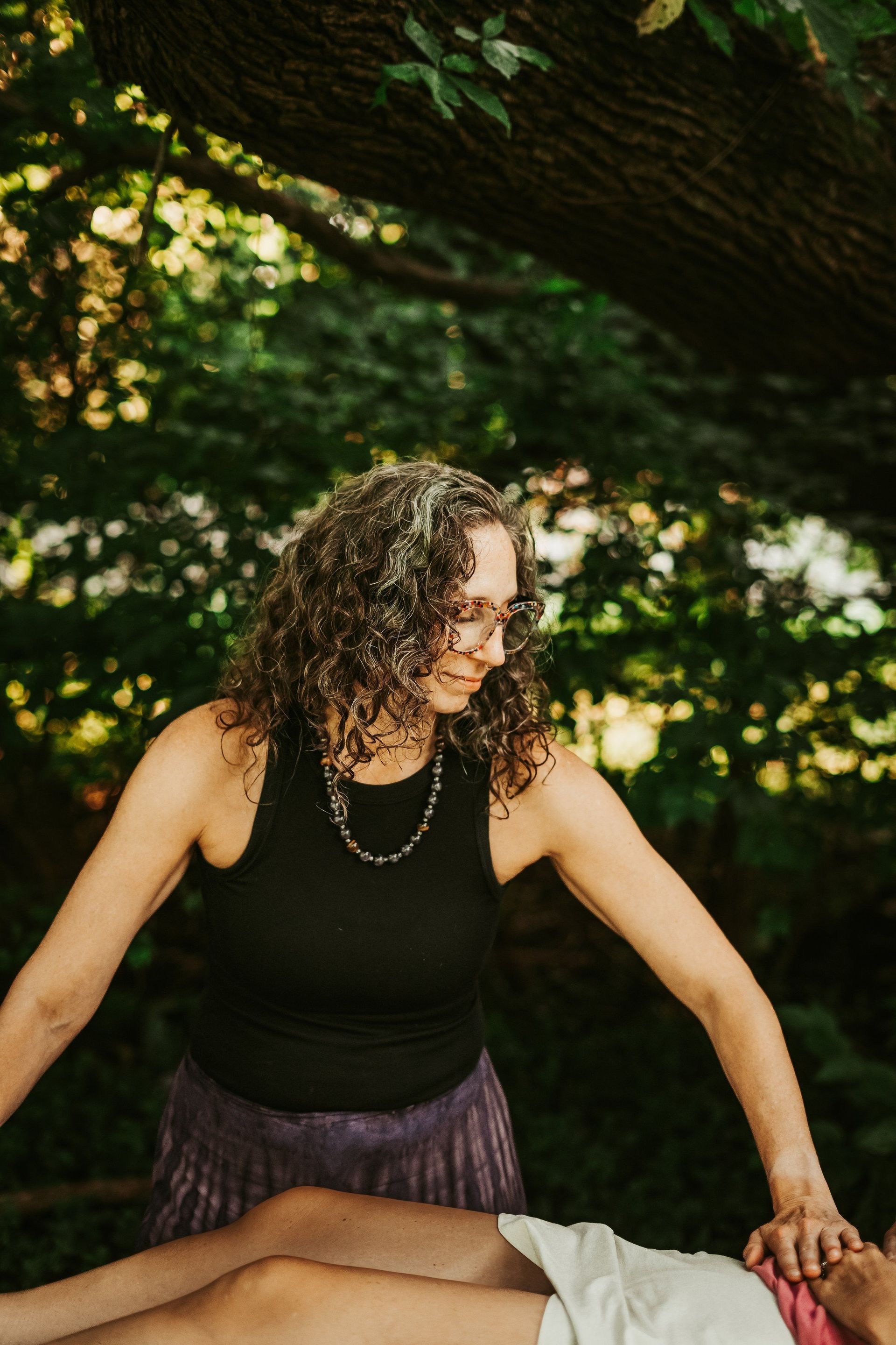 Woman with curly hair, hands over a person lying down. Outdoors, in a forested area.