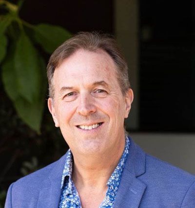Smiling man in a blue blazer and patterned shirt, standing outdoors near greenery