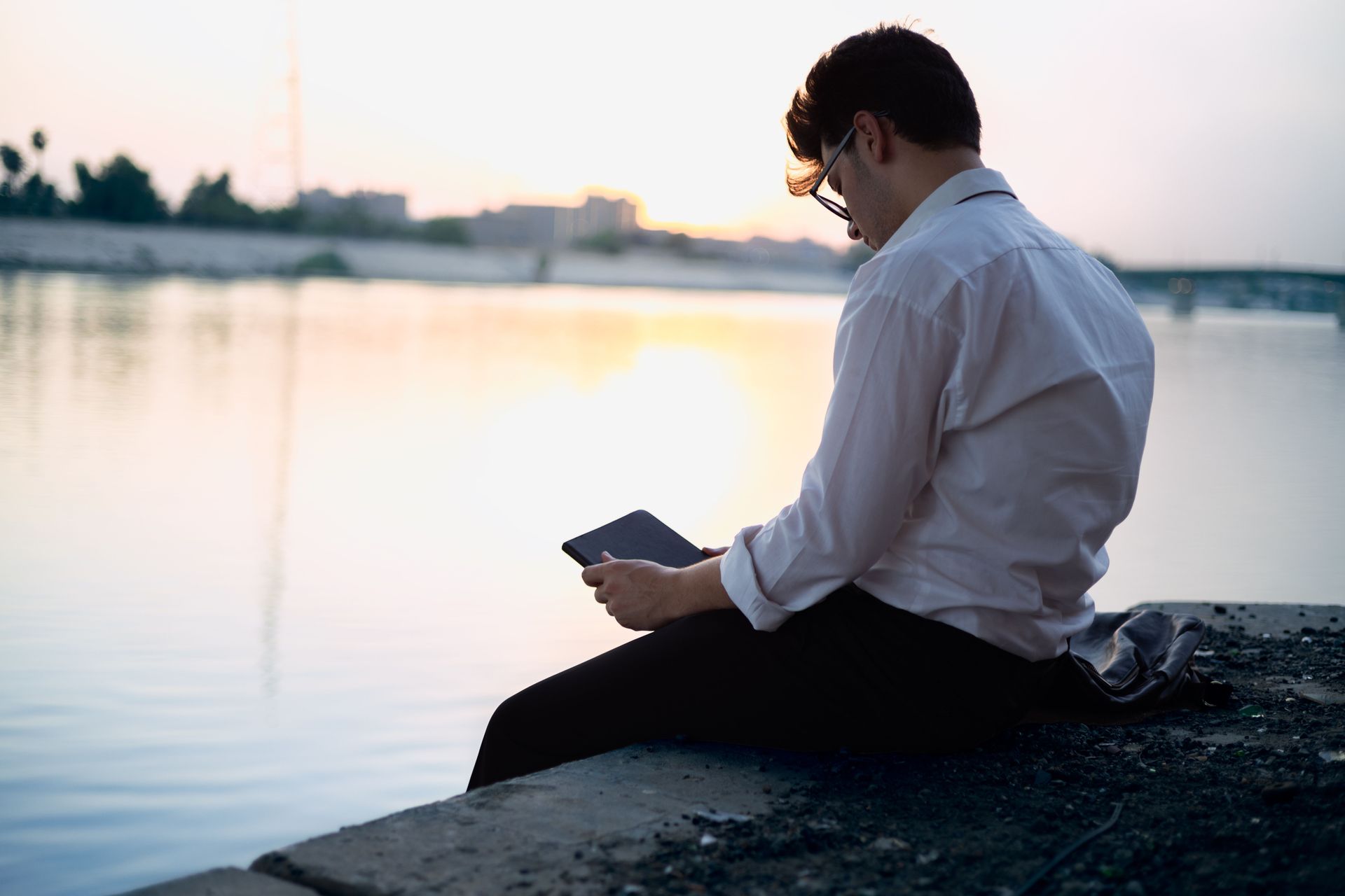 Person in a white shirt sits by calm water, looking at a phone at sunset reflecting on app strategy decision