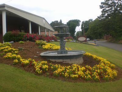 Flower beds and a stone fountain beside a building, with yellow and red flowers along a sloping lawn.