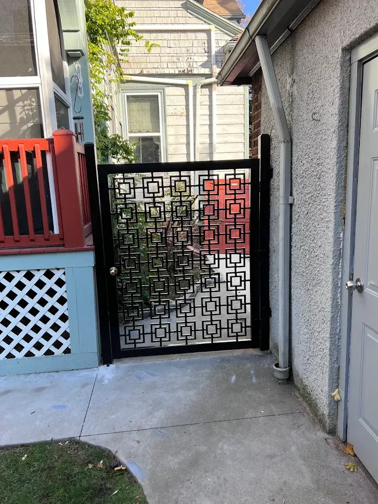 A black metal gate with a geometric square pattern stands between a house porch and a stucco wall.