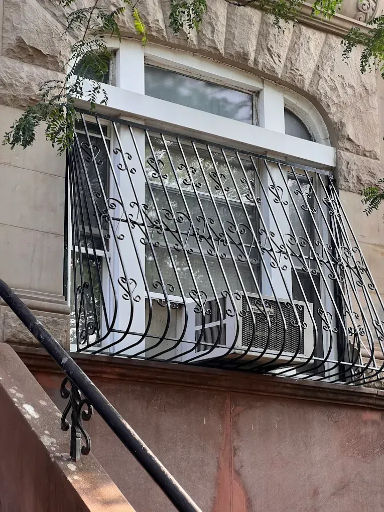 A white-framed window on a stone building, protected by a decorative black metal security grate and holding an AC unit.