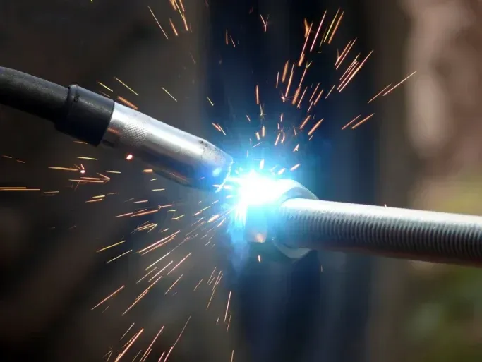 A welding torch creates a bright blue arc and flying sparks while joining metal pieces together.