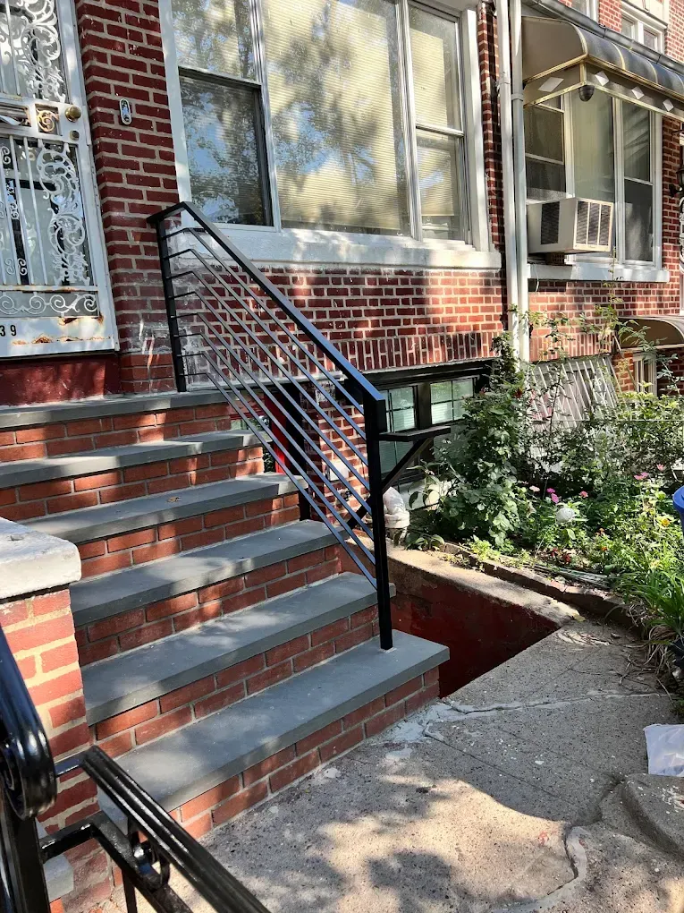 A black metal handrail with cable infill beside concrete steps leading to a brick residential building entrance.