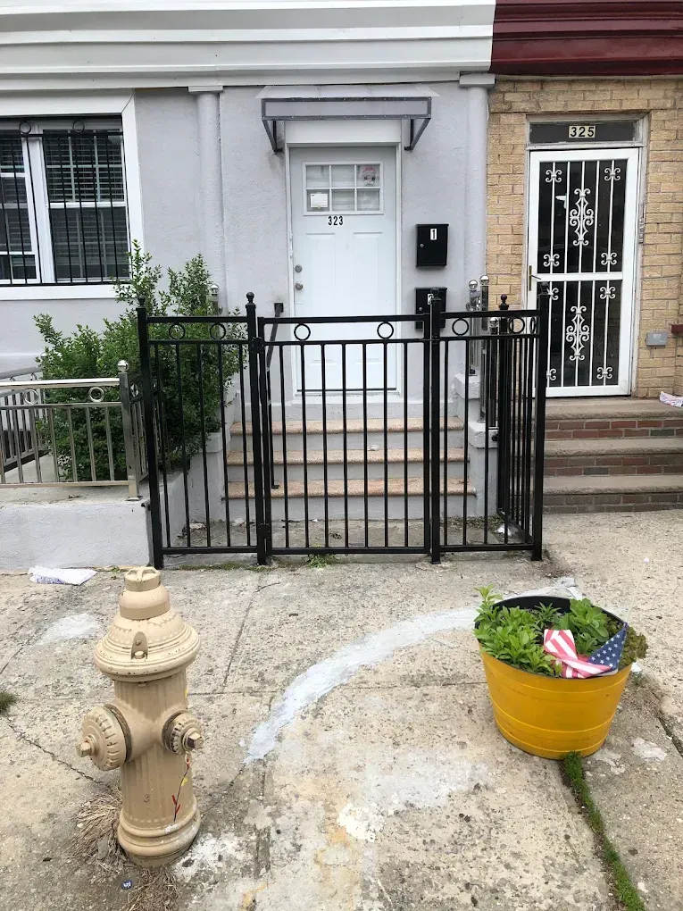 A street view of a gray row house with a black iron fence, a tan fire hydrant, and a yellow planter on the sidewalk.
