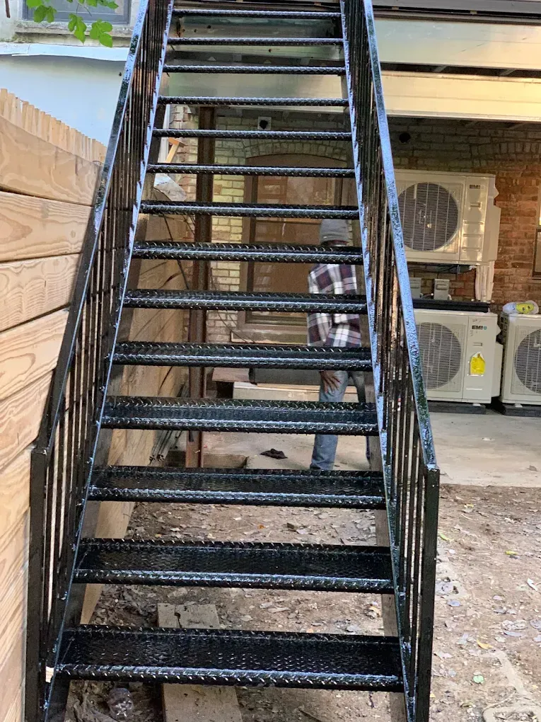 A black metal staircase leading up to an elevated deck, with outdoor air conditioning units visible underneath.
