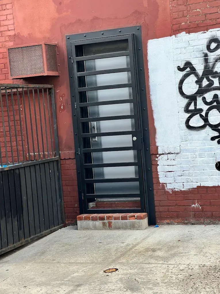 A black security gate covers a door set into a red brick wall next to graffiti and a fenced area.