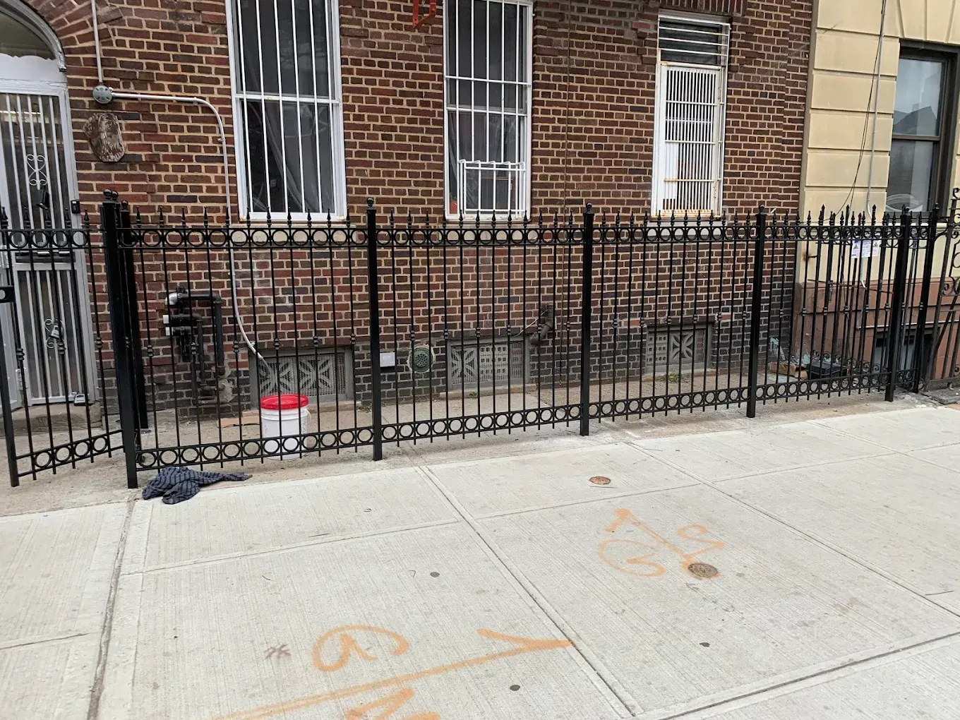 A black metal fence stands in front of a brick building with sidewalk markings in the foreground.