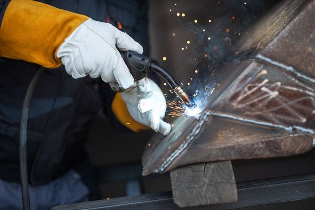 A worker wearing protective gloves uses a welding torch to join metal pieces, creating bright sparks.