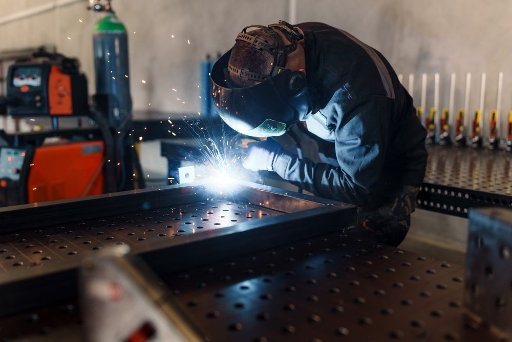 A person wearing a protective welding helmet and dark work clothes fuses metal parts on a workbench, creating sparks.