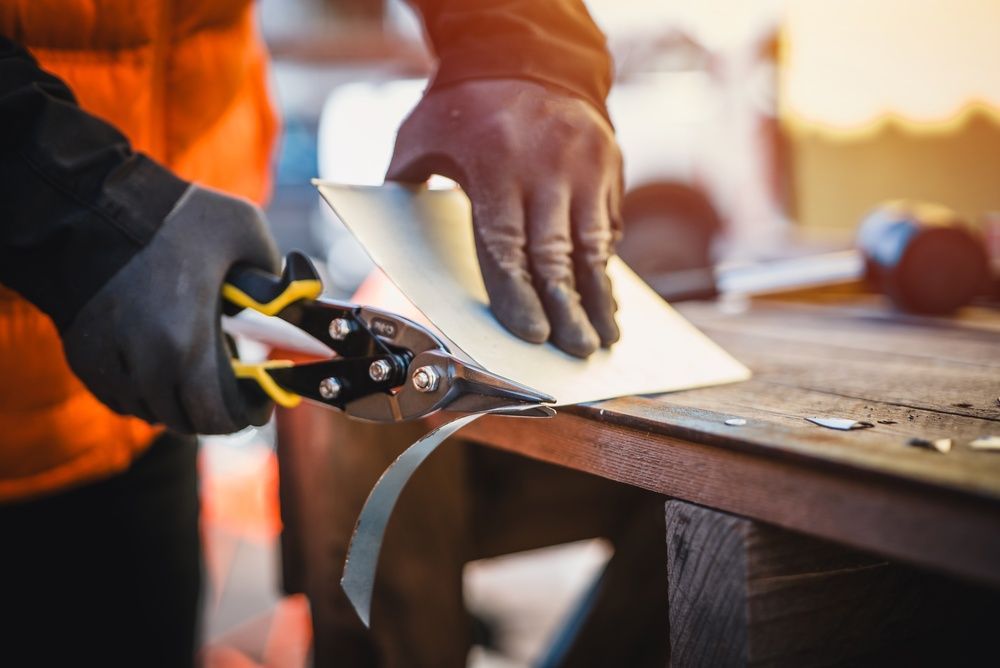 A person wearing work gloves uses metal shears to cut a sheet of metal on a wooden surface.