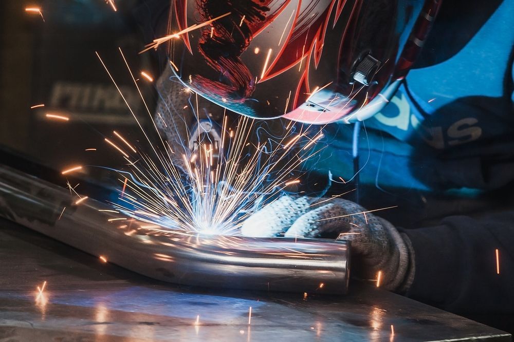A welder wearing a protective mask works on a metal pipe, creating bright sparks in a dimly lit workshop.