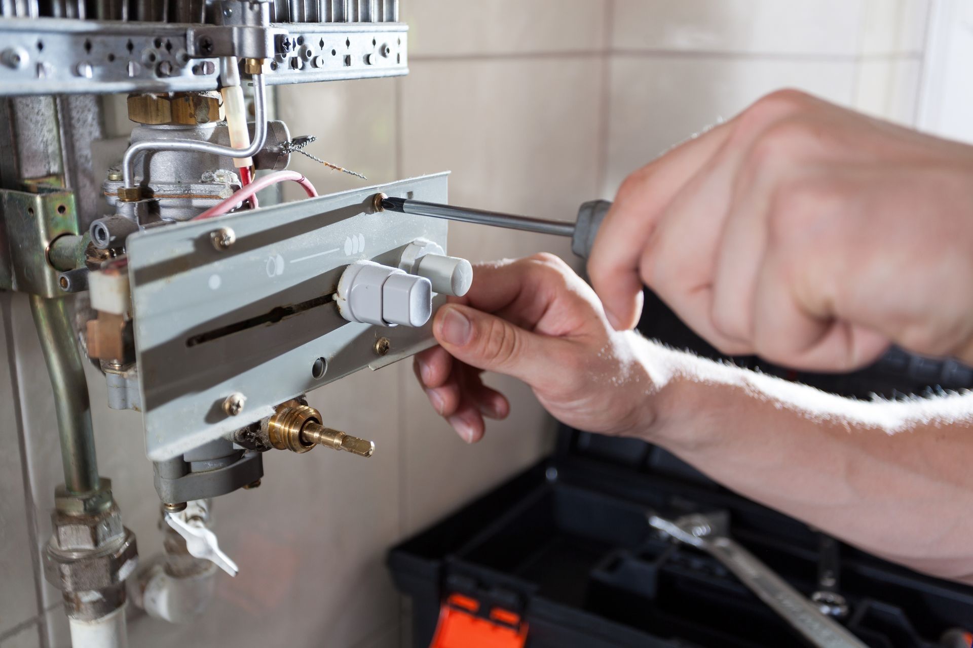 A person uses a screwdriver to work on a water heater. A toolbox sits below.