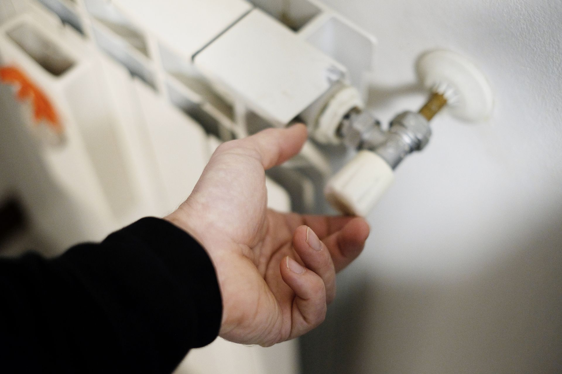 A person's hand turning a white valve on a radiator, likely adjusting the heat. The setting appears to be indoors.