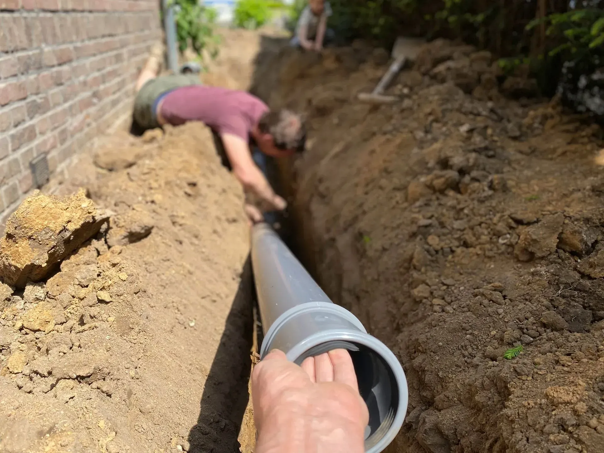 Two people install a gray pipe in a trench next to a brick wall. One person is holding the pipe, and the other is leaning into the trench.