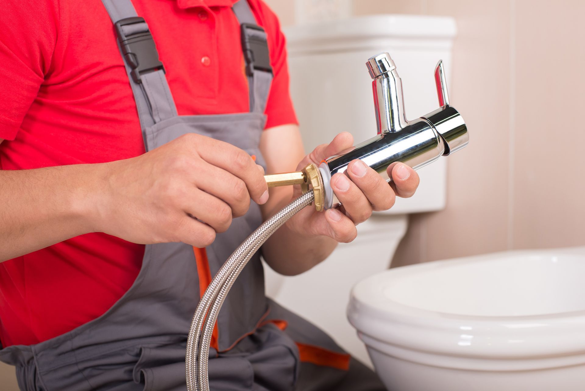 A plumber in red shirt and gray overalls attaching a faucet to a sink, near a toilet in a bathroom.