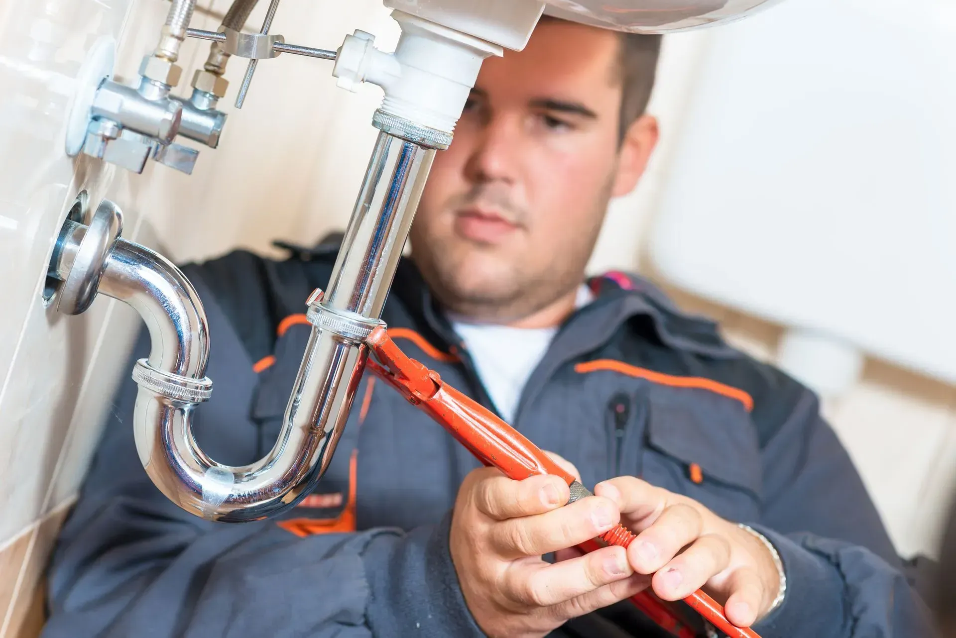 A plumber in a gray jumpsuit uses a wrench to work on a sink's plumbing. He is focused and in a bathroom setting.