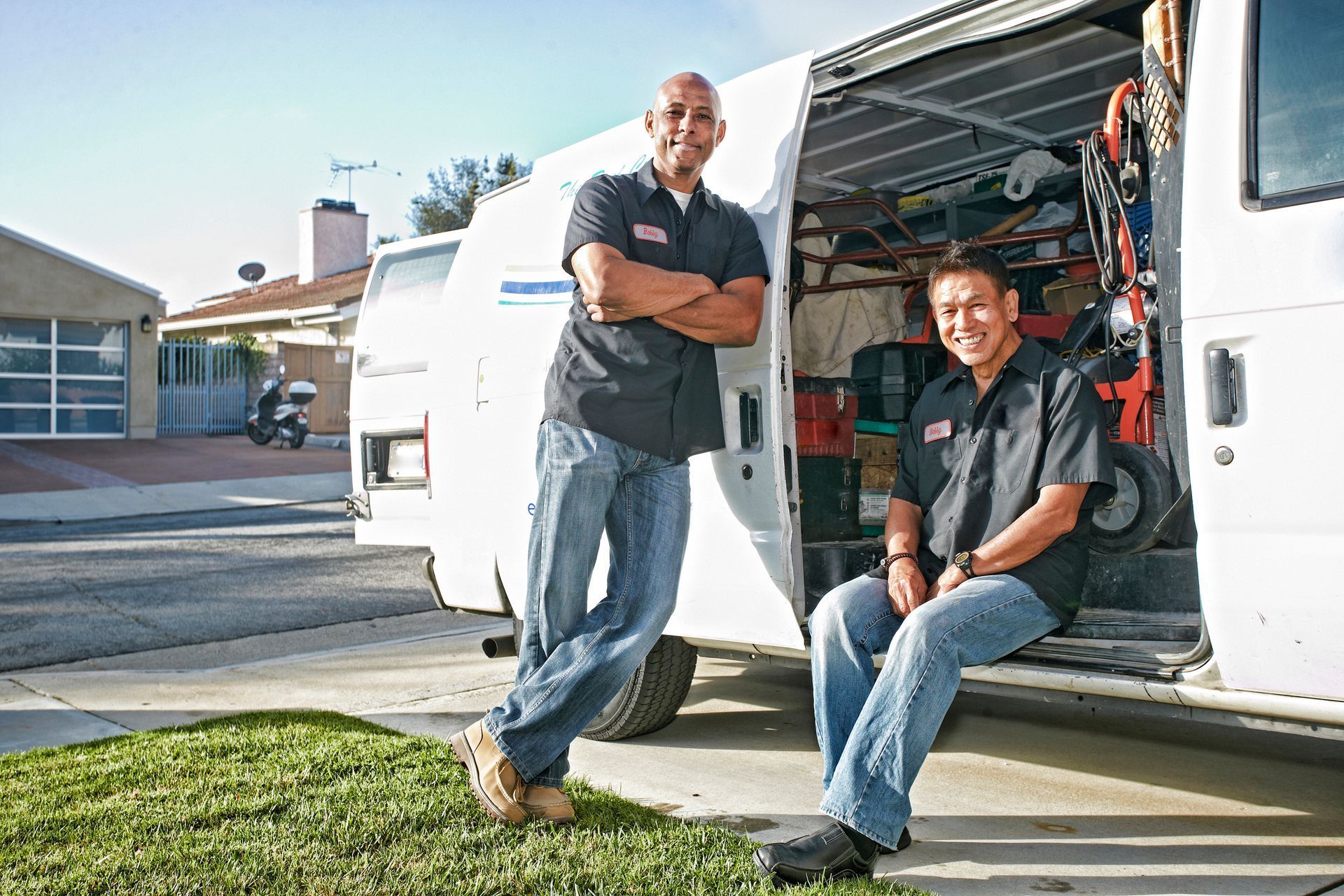 Two men standing by the open back of a white service van parked on a lawn. Both wear blue jeans and short-sleeved shirts; one has arms crossed.
