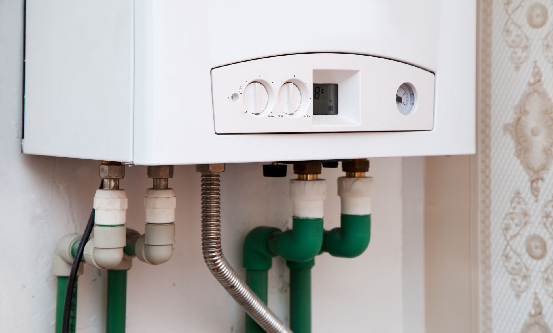 White wall-mounted boiler with dials and green pipes, set against a patterned wallpaper.