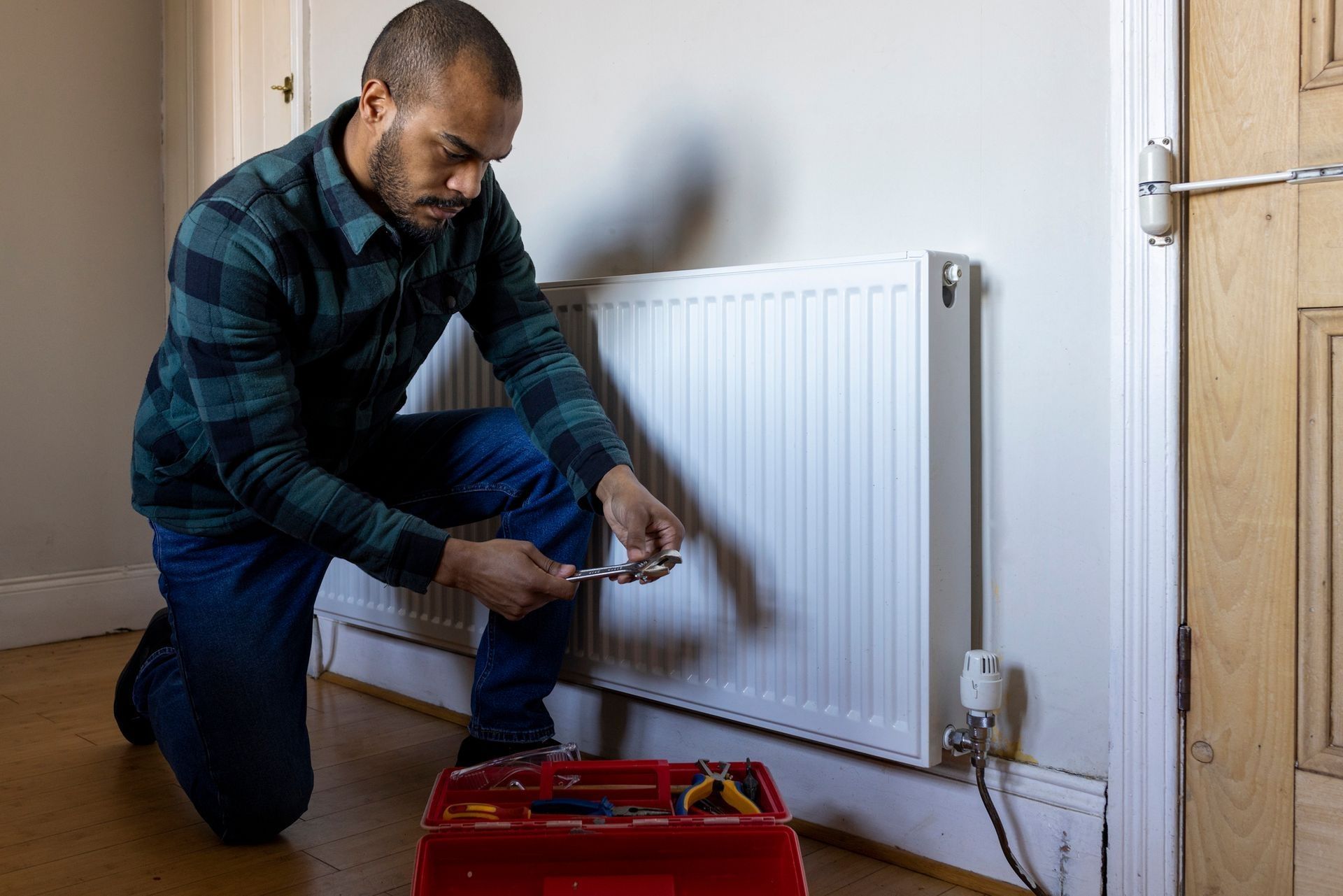 Man kneeling, repairing a white radiator with tools from a red toolbox in a room. He wears a plaid shirt and jeans. Man kneeling, repairing a white radiator with tools from a red toolbox in a room. He wears a plaid shirt and jeans.