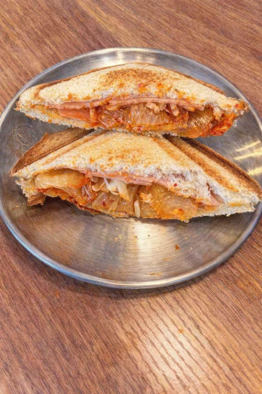 Two triangular kimchi and ham toasted sandwiches served on a silver plate against a wooden background.