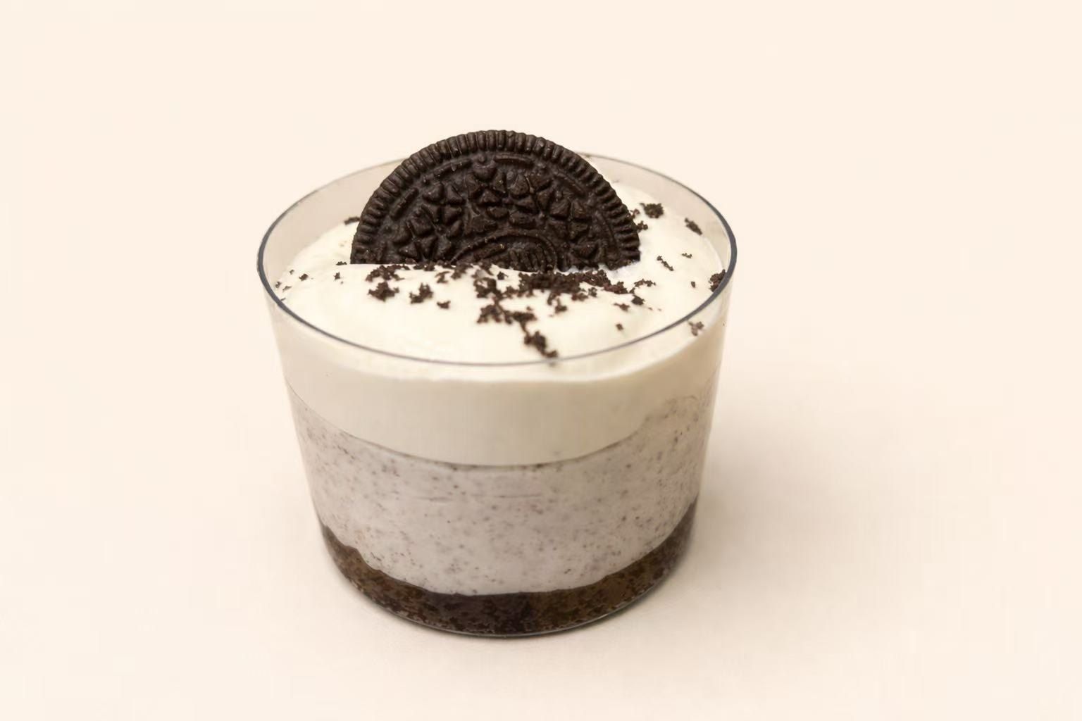 A layered Oreo dessert in a clear plastic cup, topped with a whole cookie and crushed cookie pieces on white cream.