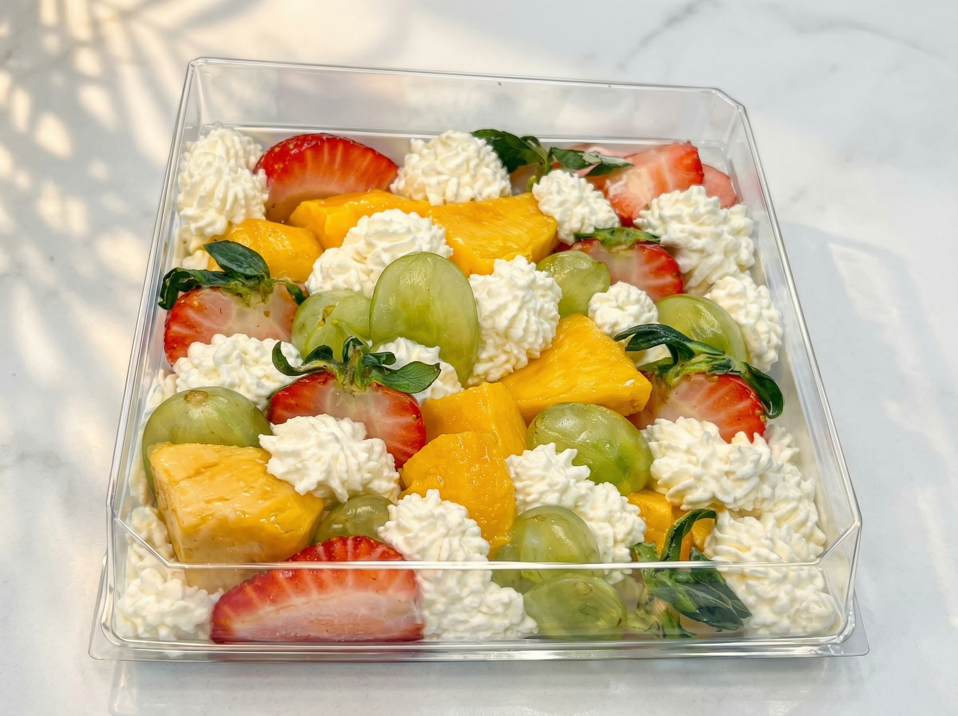 A clear square container filled with a colorful fruit salad of mango, grapes, and strawberries topped with whipped cream.