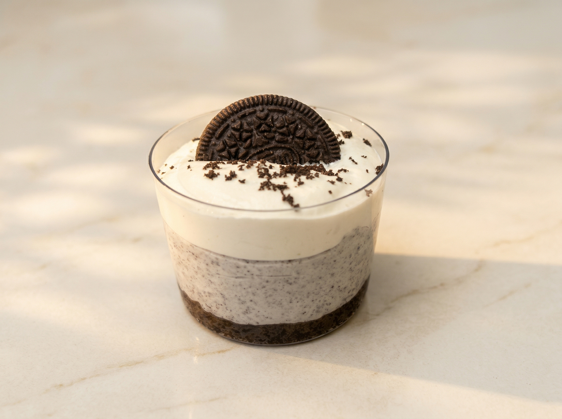 A layered Oreo dessert in a clear plastic cup, topped with a whole Oreo cookie, sitting on a light-colored surface.