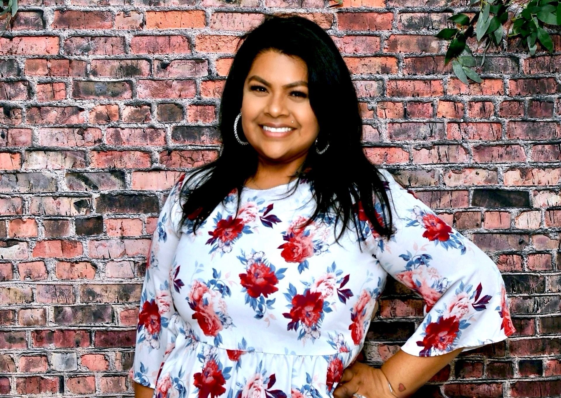 A woman in a floral dress is standing in front of a brick wall.