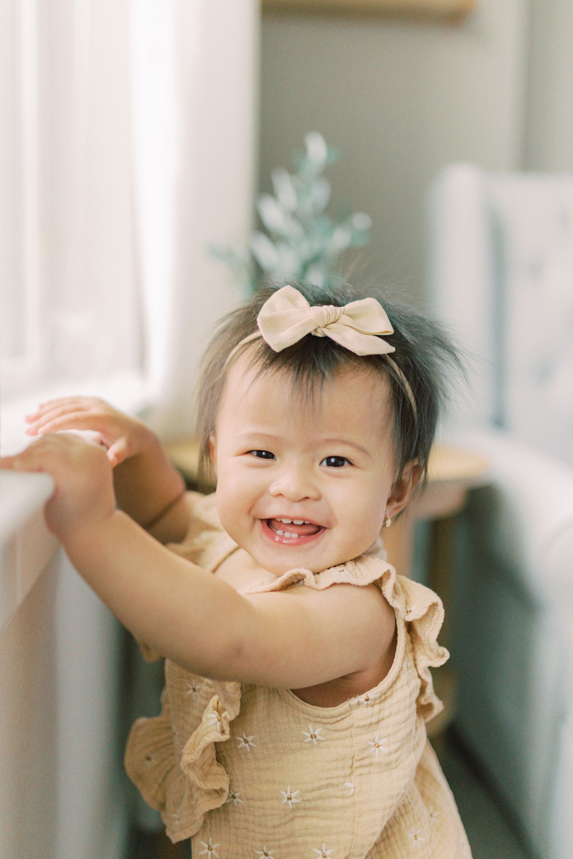 Smiling baby, standing at a window, wearing a bow and tan romper, indoors.