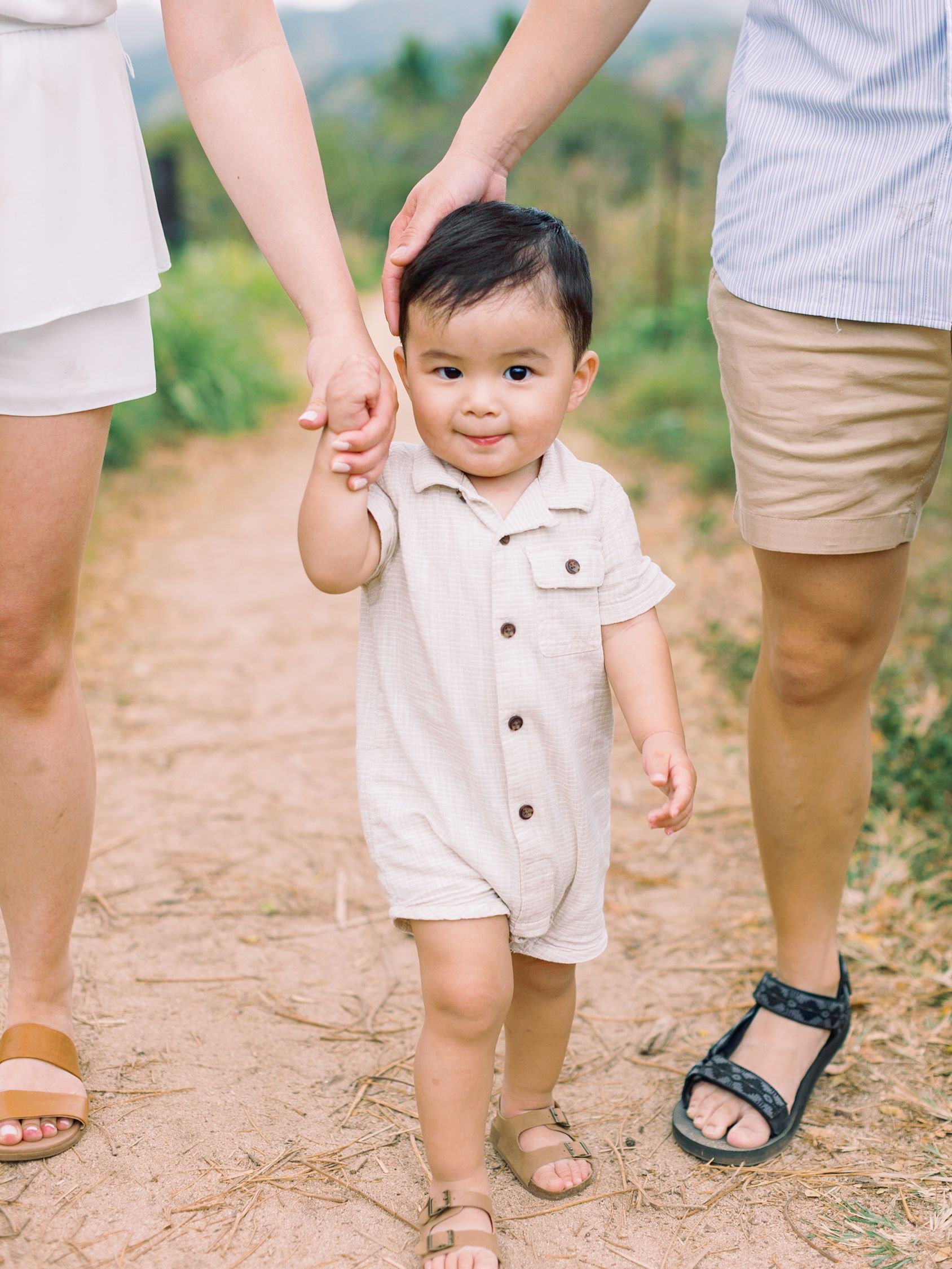 Toddler walking outdoors, holding hands with two adults on a sandy path