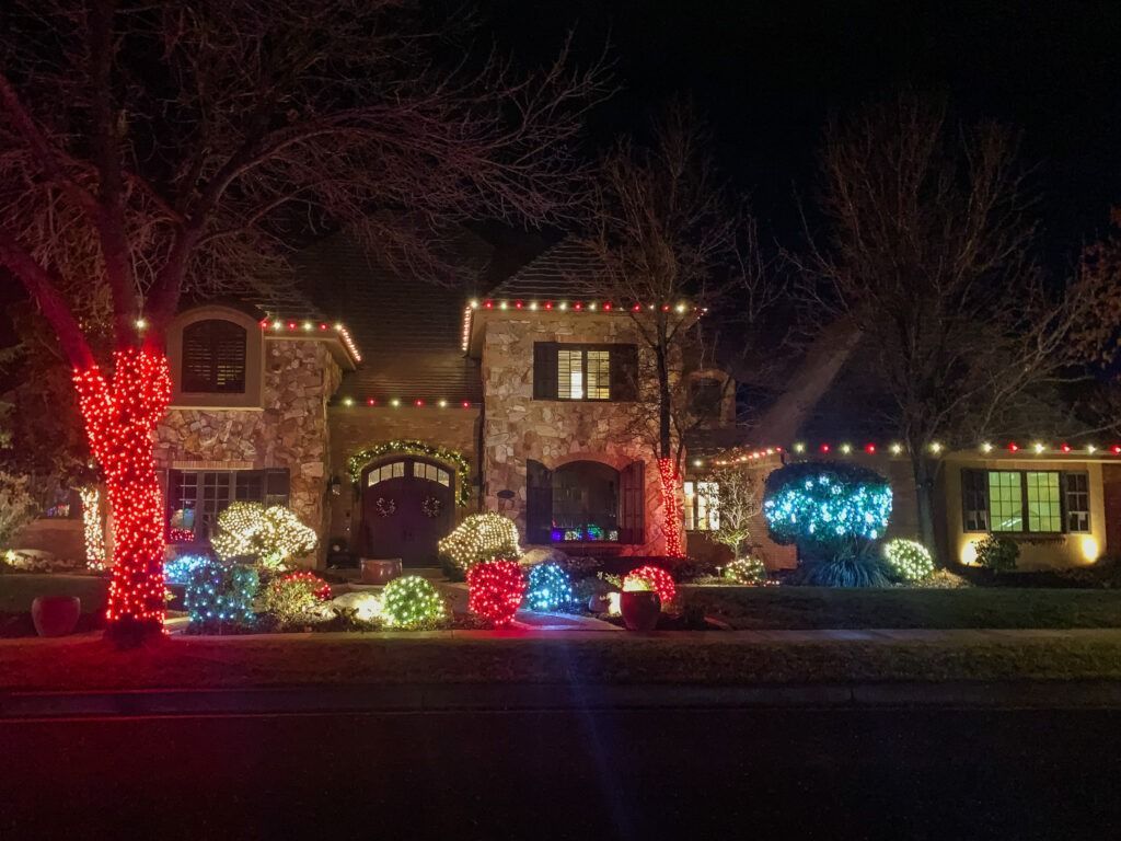 A multi-story house decorated for the holidays with bright, colorful outdoor lights, glowing bushes, and a lit-up tree.