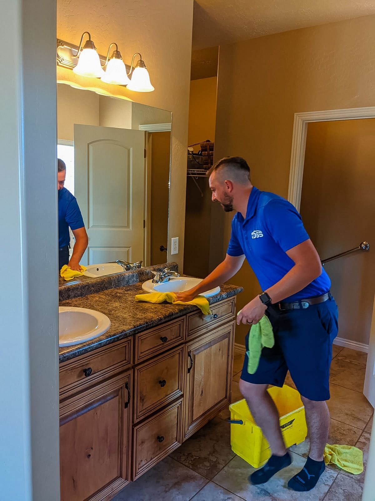 A person in a blue uniform cleaning a bathroom countertop with a yellow cloth, reflected in a vanity mirror.