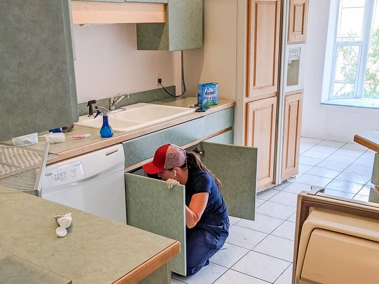 A person in a red cap is kneeling and working inside an open cabinet under a kitchen sink.