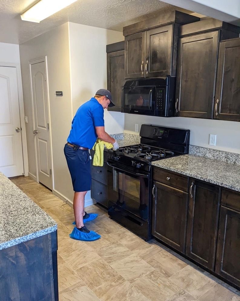 A person in a blue shirt and protective shoe covers cleans a black stove in a kitchen with dark wood cabinets.