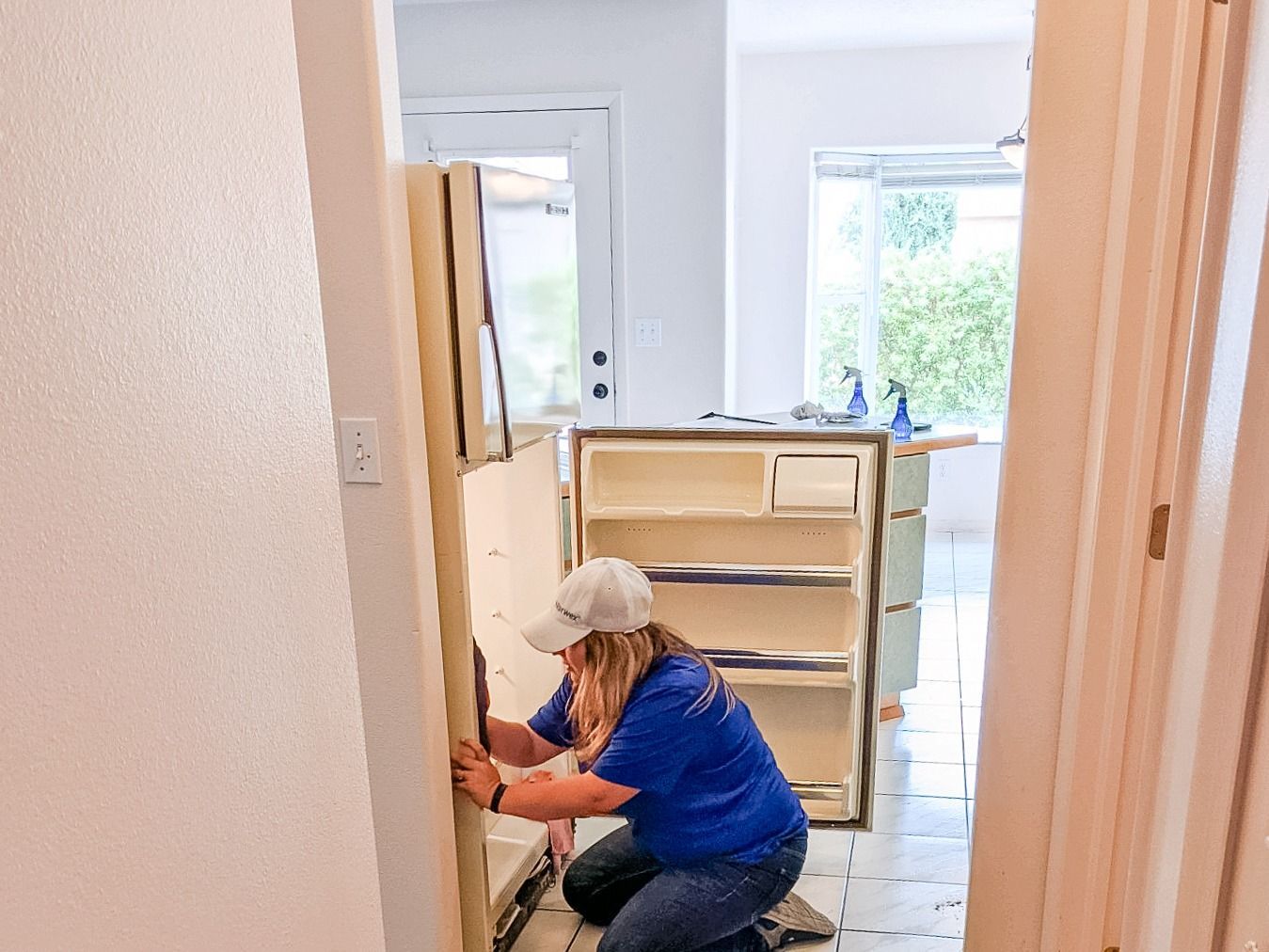 A person in a blue shirt kneels on a kitchen floor, inspecting the inside of an open, empty refrigerator.