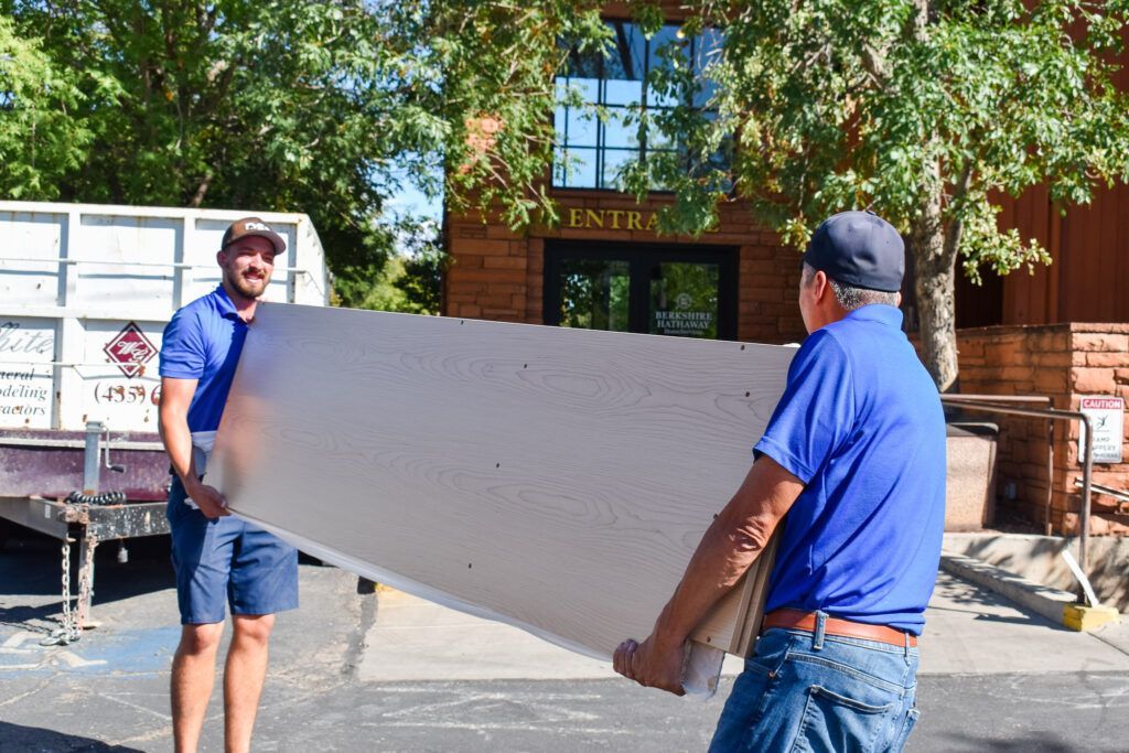 Two people in blue shirts carry a large rectangular construction panel outdoors near a trailer and building entrance.
