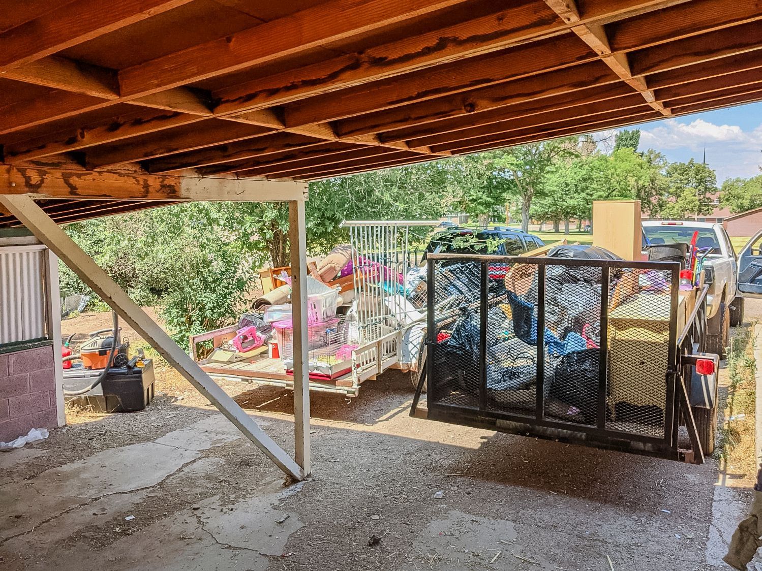 A truck parked under a wooden patio cover is loaded with a cluttered trailer full of items and miscellaneous household junk.