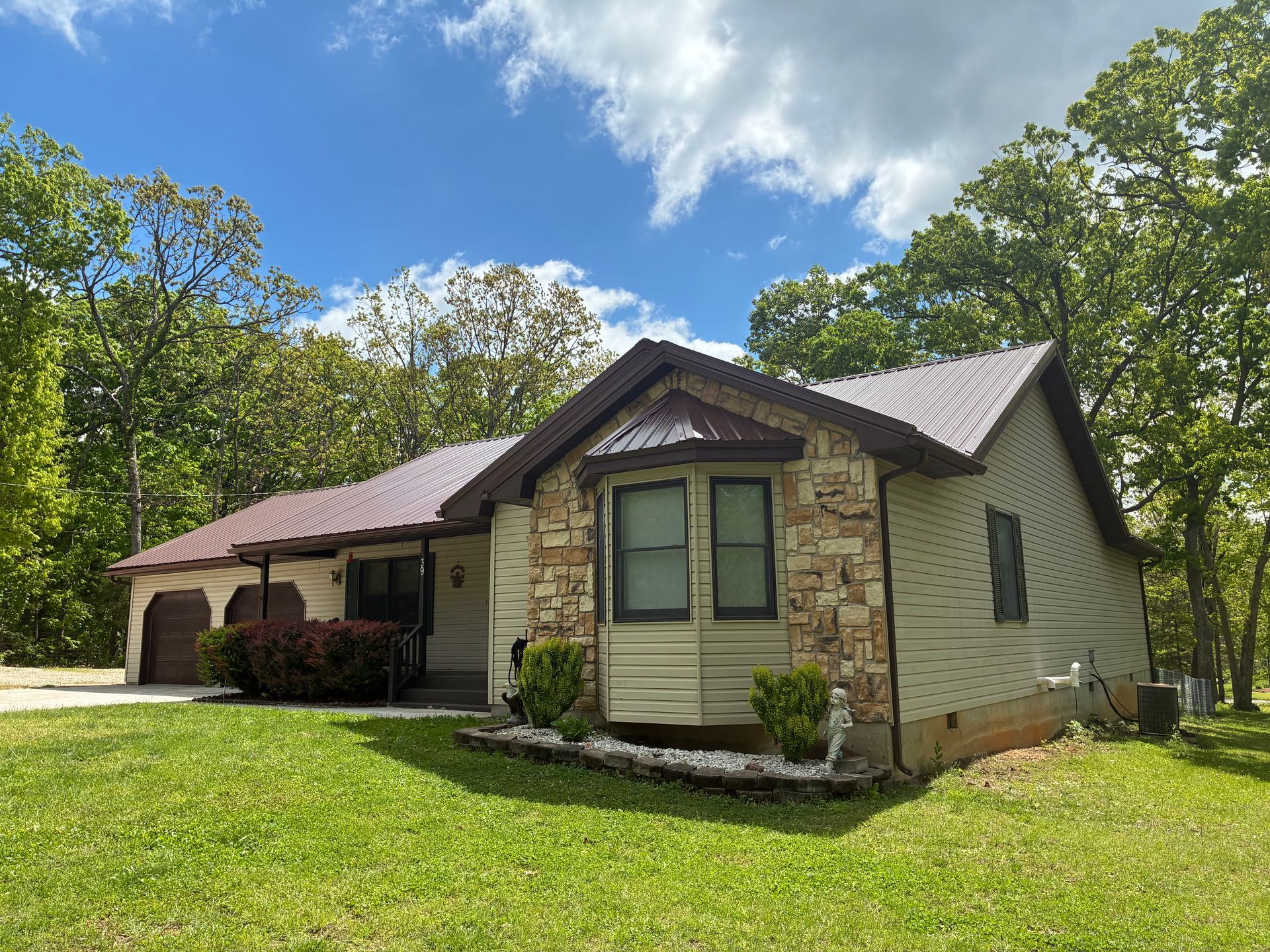 House with brown metal roof, stone facade, green lawn, and surrounding trees.