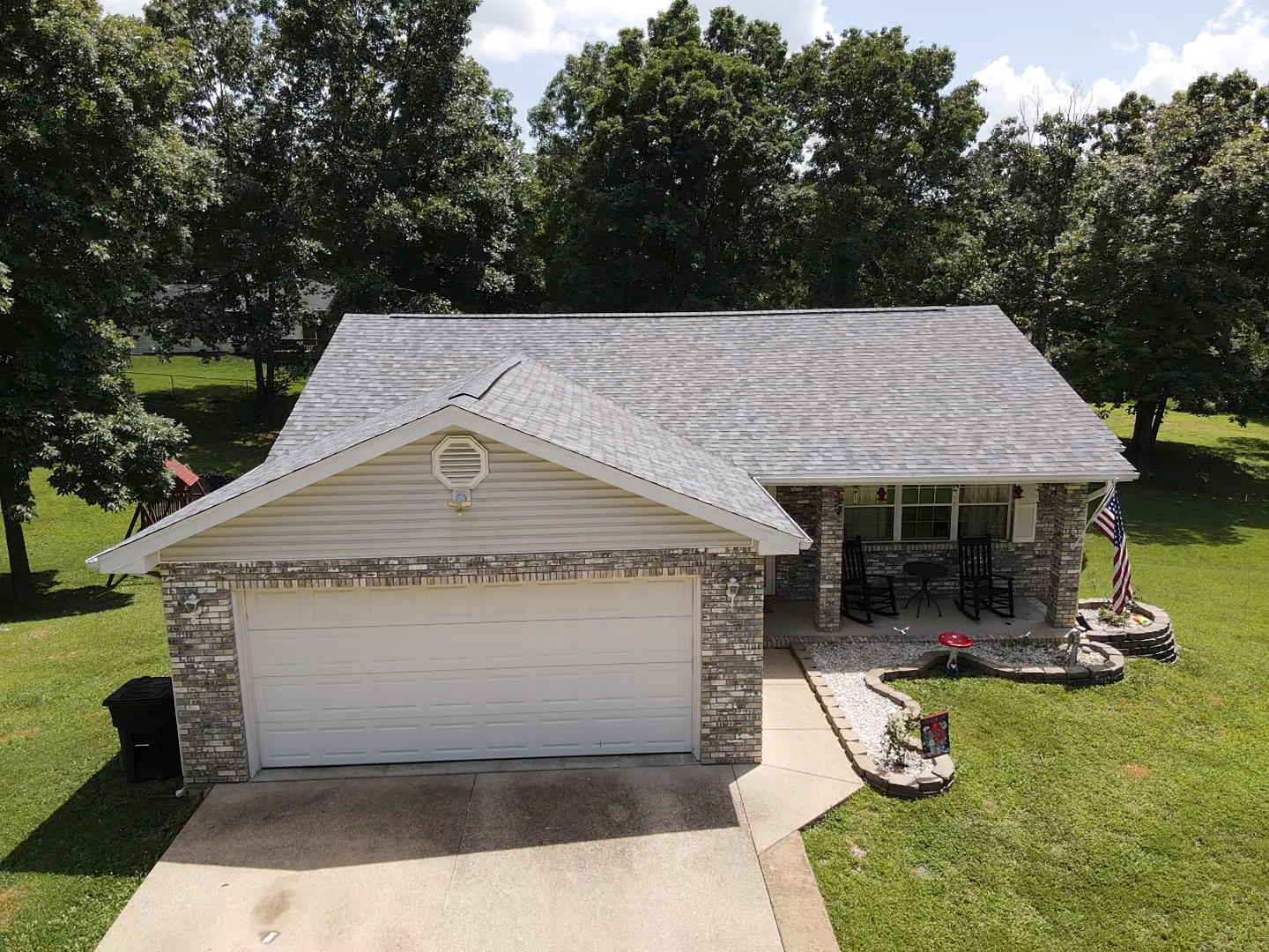 House with gray roof, tan siding, and stone accents. Features a garage, porch, and American flag in a grassy yard.