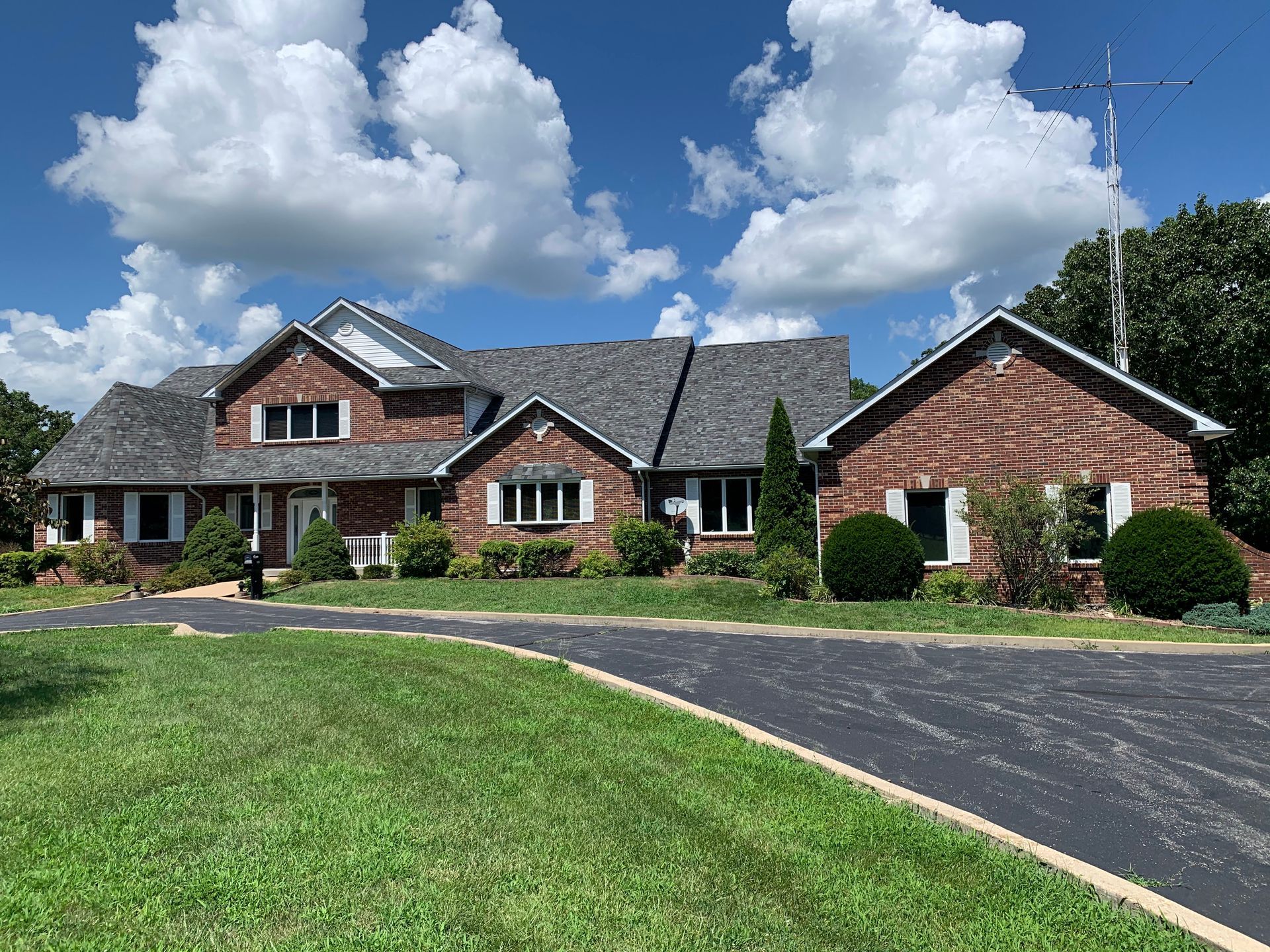 Brick house with asphalt driveway on a sunny day. Green lawn, white trim, and fluffy clouds.